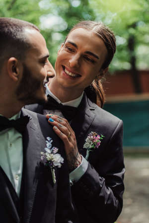 happy gay newlyweds in suits with boutonnieres looking at each otherの写真素材