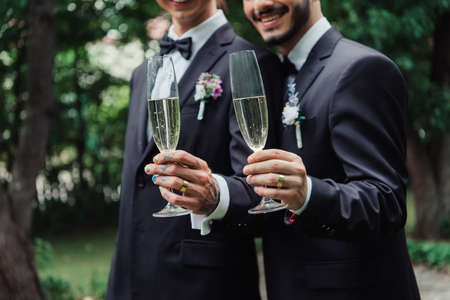 cropped view of cheerful gay newlyweds in suits holding glasses with champagne on wedding dayの写真素材