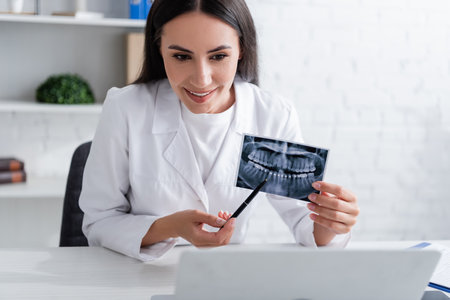 Smiling doctor pointing at scan of teeth near laptop in clinicの写真素材