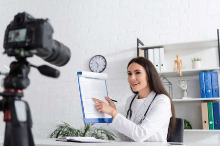 Smiling doctor pointing at clipboard near blurred digital camera in hospital
