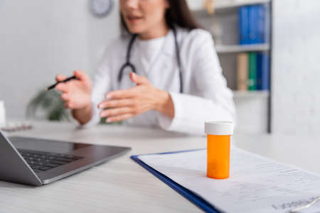 Cropped view of pills on clipboard near blurred doctor having video call on laptop in clinicの写真素材