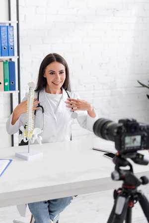Smiling doctor looking at digital camera near spinal model and smartphone in clinicの写真素材