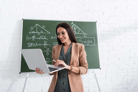Smiling teacher using laptop near math formulas on chalkboard in school