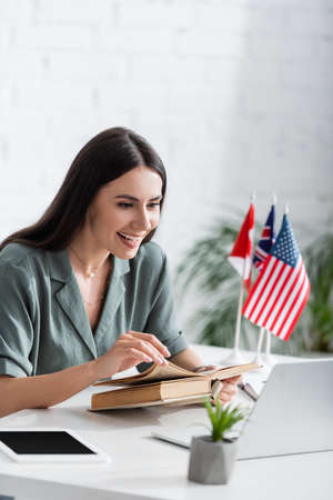 Cheerful teacher holding book and looking at laptop during online lecture in schoolの写真素材