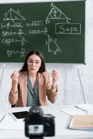 Teacher talking near blurred digital camera and chalkboard with math formulas in classroomの写真素材