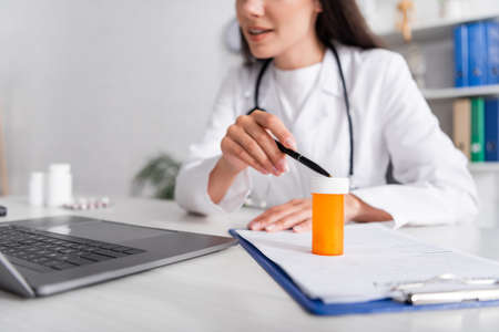 Cropped view of doctor pointing at jar with pills on clipboard during online consultation on laptop in clinicの写真素材