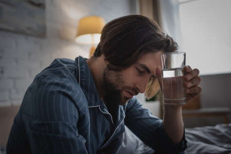 Tired man in pajama holding glass of water in bed in morningの写真素材