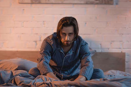 Upset man in pajama looking at camera while suffering from insomnia on bed
