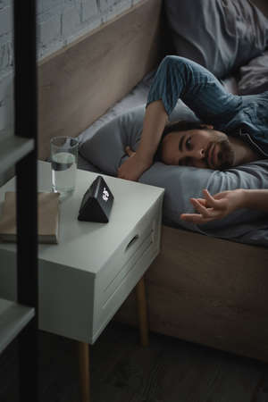 Man with insomnia looking at alarm clock near book and water on bedside table at home