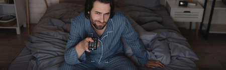 Dissatisfied man in pajama clicking channels while sitting on bed at night, bannerの写真素材