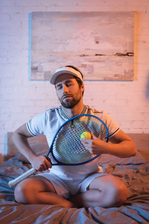Noctambulist in sportswear holding tennis ball and rocket on bed at nightの写真素材