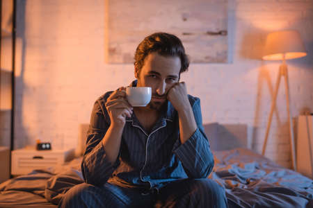 Displeased man with sleep disorder holding cup and looking at camera on bedの写真素材