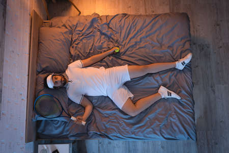 Top view of man in sportswear holding racket and tennis ball while sleeping on bedの写真素材
