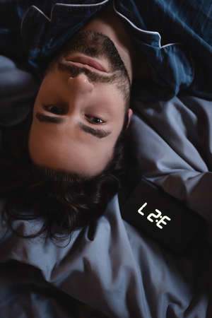 Top view of young man in pajama looking at camera near alarm clock on bedの写真素材