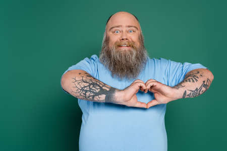 happy plus size man with beard and tattoos showing heart sign isolated on greenの写真素材