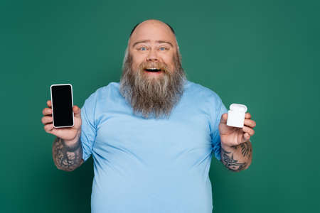 cheerful plus size man with mobile phone and wireless earphones isolated on green
