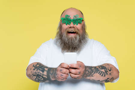 amazed plus size man in clover-shaped glasses chatting on smartphone isolated on yellow