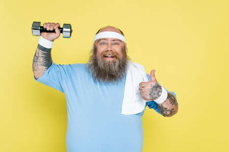 cheerful plus size man showing thumb up while exercising with dumbbell isolated on yellow