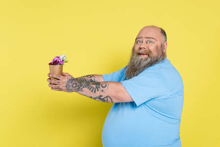 happy plus size man with beard holding paper cup with flowers isolated on yellow