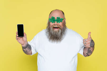 joyful overweight man in clover-shaped glasses showing thumb up while holding smartphone isolated on yellowの写真素材