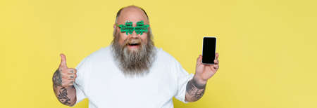 excited plus size man in clover-shaped glasses holding smartphone and showing thumb up isolated on yellow, bannerの写真素材