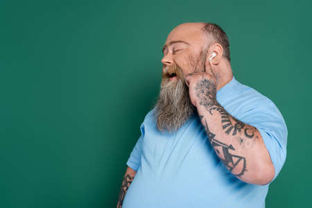 pleased overweight man with closed eyes listening music in earphone and singing isolated on green