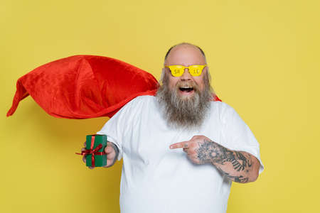 amazed plus size man in glasses with sale lettering and red cloak pointing at present isolated on yellowの写真素材
