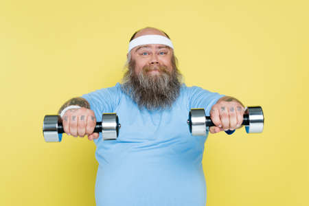 chubby and bearded man working out with dumbbells and smiling at camera isolated on yellow