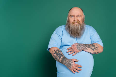 displeased overweight man hugging tummy while suffering from stomach pain isolated on greenの写真素材