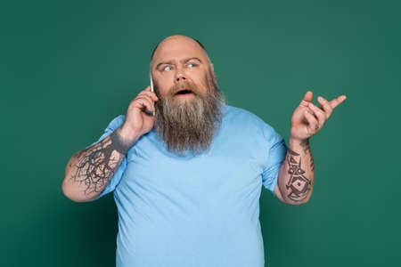 thoughtful overweight man with tattoos talking on smartphone and pointing with finger isolated on greenの写真素材