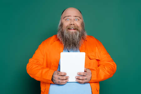 amazed and happy plus size man with beard holding digital tablet isolated on greenの写真素材