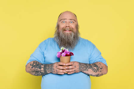 excited plus size man with tattoos holding paper cup with colorful flowers isolated on yellow