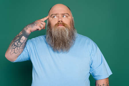 tattooed plus size man touching head and looking away while thinking isolated on green