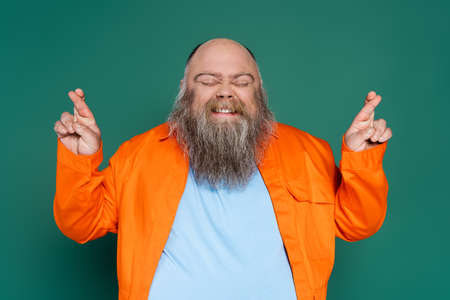tense bearded man with closed eyes holding crossed fingers for luck isolated on green