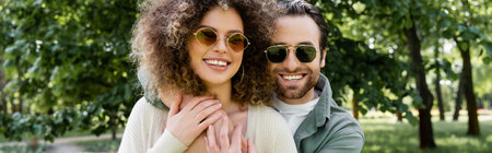 happy man in stylish sunglasses hugging curly girlfriend in park, banner