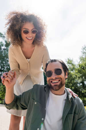 cheerful man and pleased curly woman in trendy sunglasses smiling in parkの写真素材