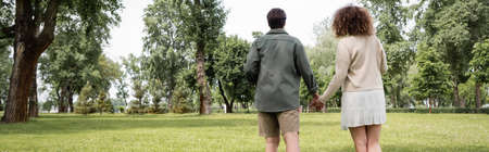 back view of curly woman in dress and man in summer clothes walking together in park, banner