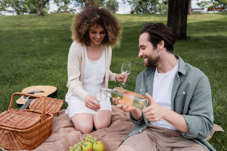 cheerful man pouring wine in glass near curly woman during summer picnic