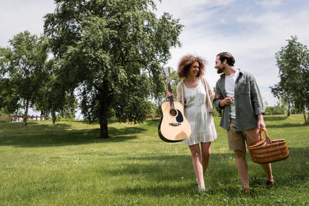 full length of smiling curly woman holding acoustic guitar near boyfriend with wicker basket walking in parkの写真素材