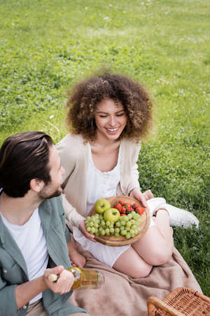 high angle view of happy man holding bottle of wine near curly girlfriend sitting with cutting board and fruitsの写真素材
