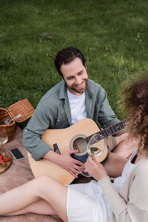 high angle view of romantic man playing acoustic guitar near curly woman with glass of wine during...