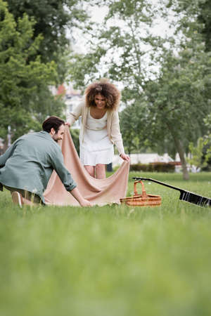happy young couple in summer clothes putting picnic blanket on green grass in parkの写真素材