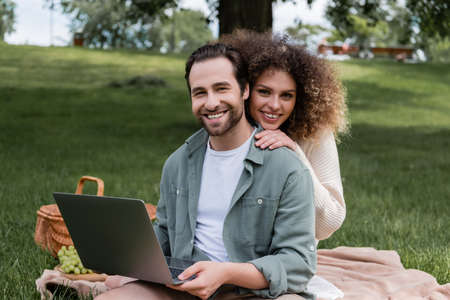 cheerful man holding laptop and smiling with curly woman during picnicの写真素材