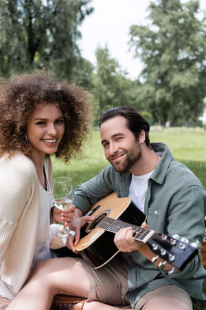 romantic man playing acoustic guitar near smiling woman with glass of wine during picnicの写真素材
