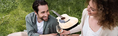 high angle view of happy curly woman passing glass of wine to boyfriend during summer picnic, banner