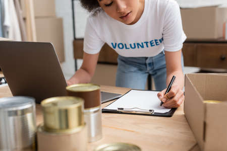 cropped view of african american volunteer writing on clipboard near laptop and blurred tinsの写真素材