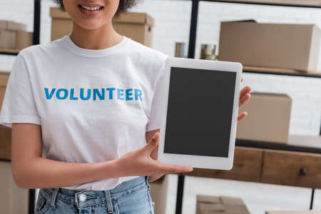 partial view of smiling african american volunteer holding digital tablet with blank screen in...