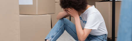tired african american volunteer obscuring face with hands near carton boxes in donation center, bannerの写真素材