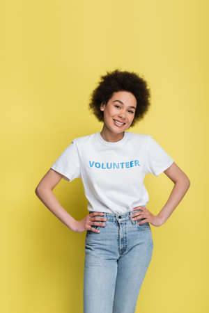 pleased african american volunteer posing with hands on hips isolated on yellowの写真素材