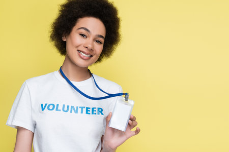 african american volunteer smiling at camera and showing blank id card isolated on yellowの写真素材